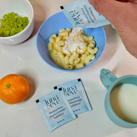 Bowl of macaroni and cheese with a hand to sprinkle Luna & Eve flavourless multivitamin powder packet, surrounded by an orange and a cup of milk.