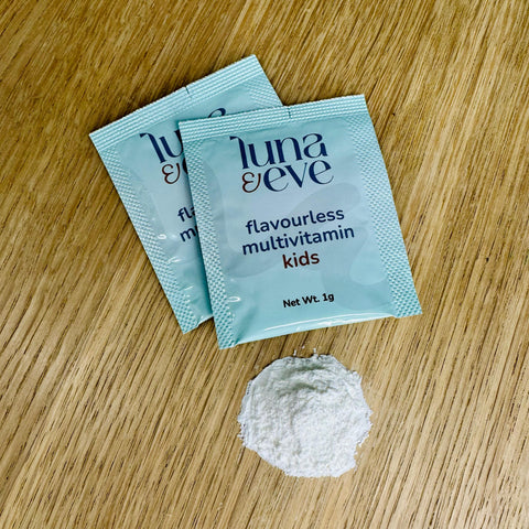 Two packets of Luna & Eve flavourless multivitamin kids on a wooden surface with powder within one sachet beside them.